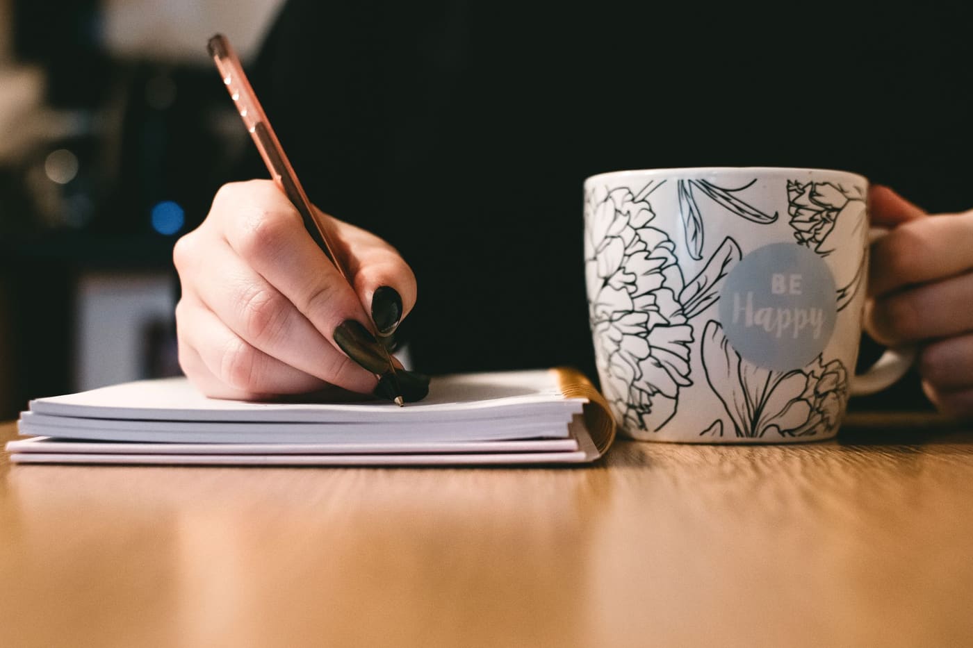 A person writing in a notebook beside a floral mug on a wooden desk