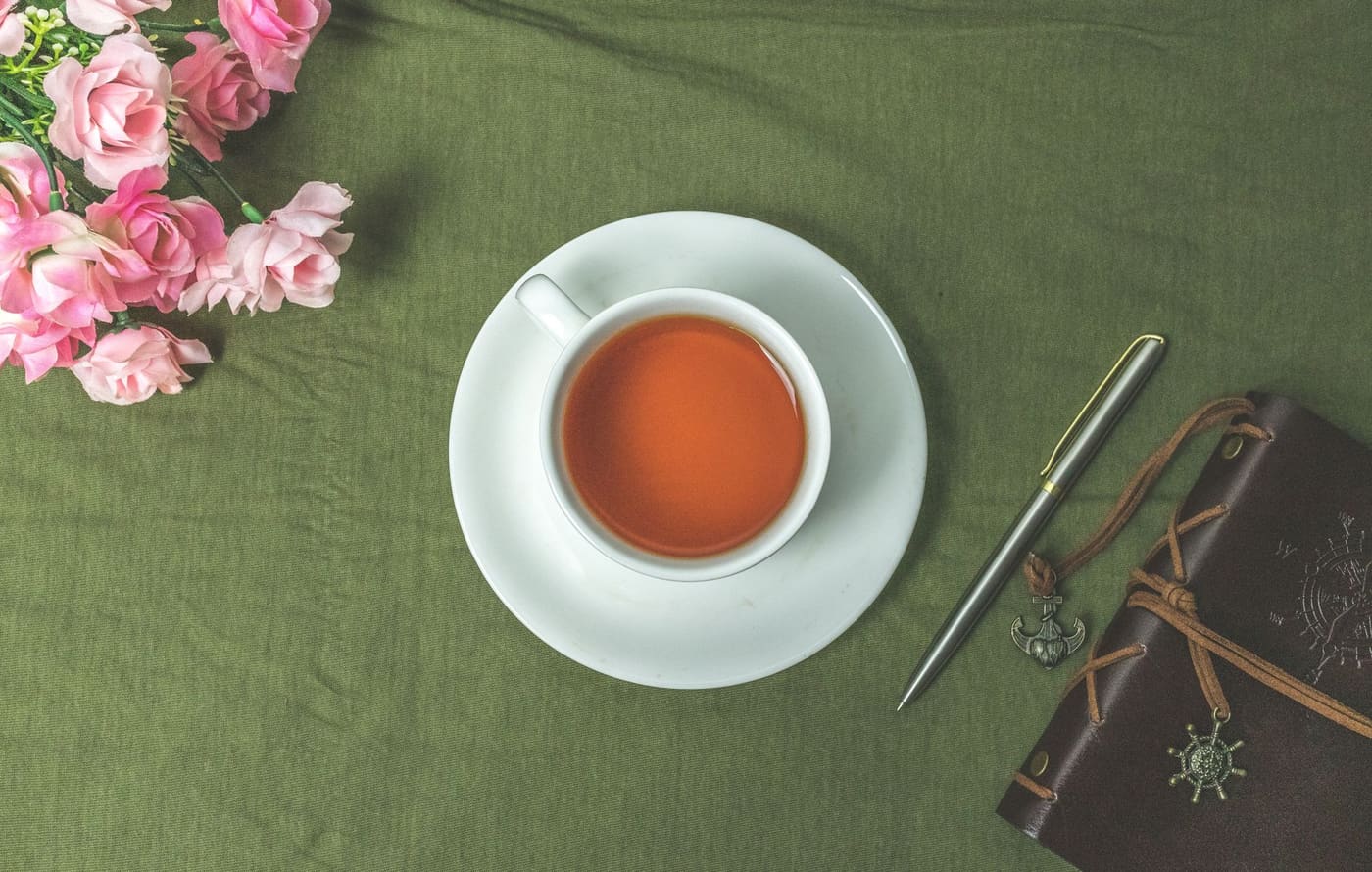 A cup of tea, pink flowers, a pen and a leather journal on a green cloth