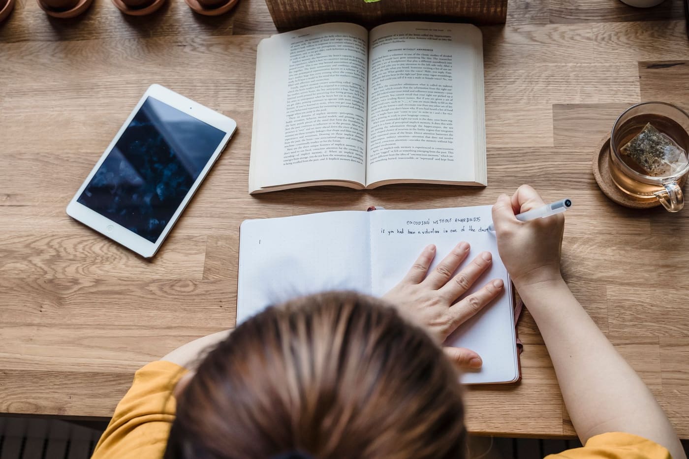 Overhead view of a person writing in a notebook on a wooden table with tea and a tablet