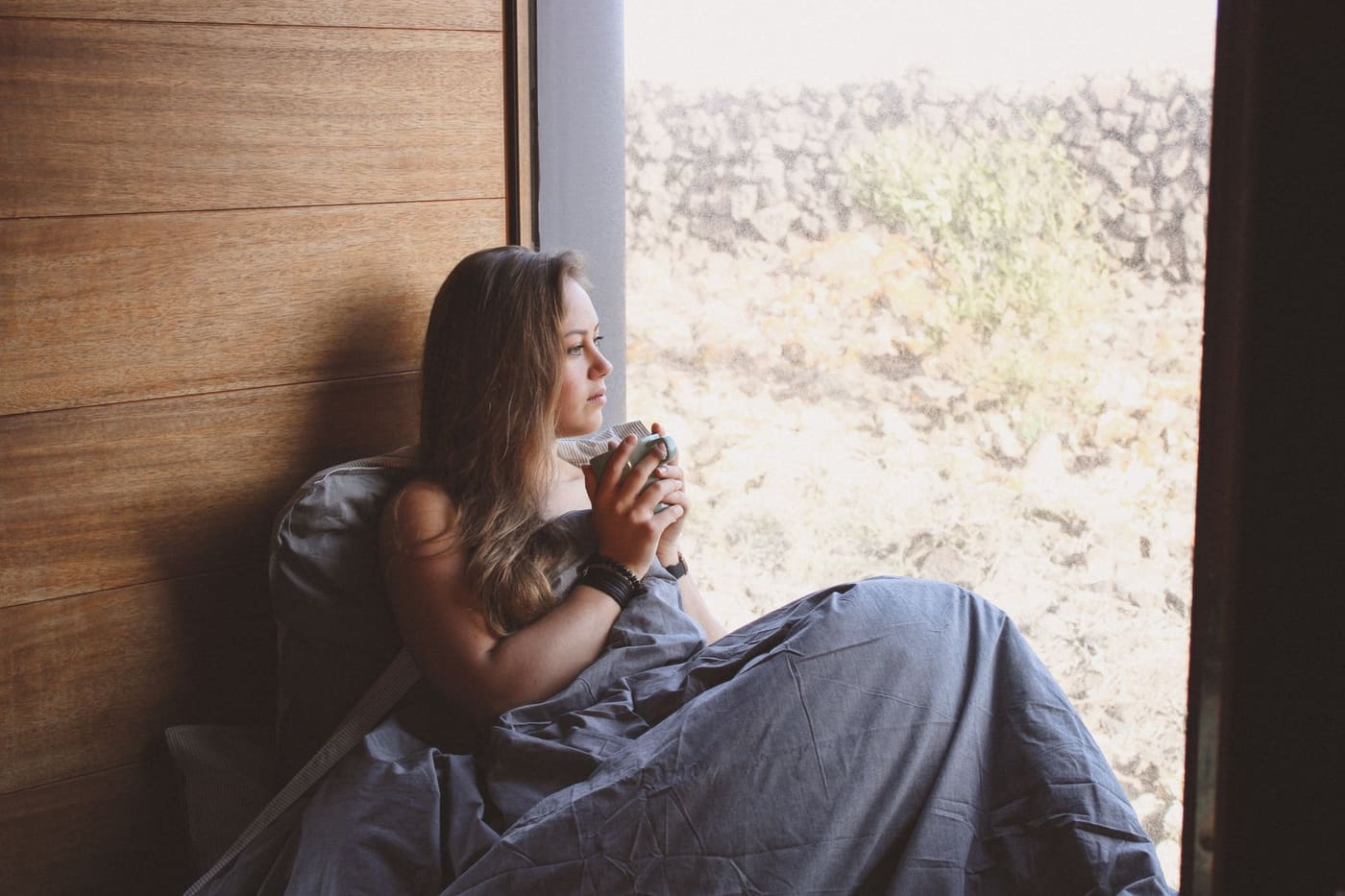 A woman wrapped in a blanket by a window holding a mug