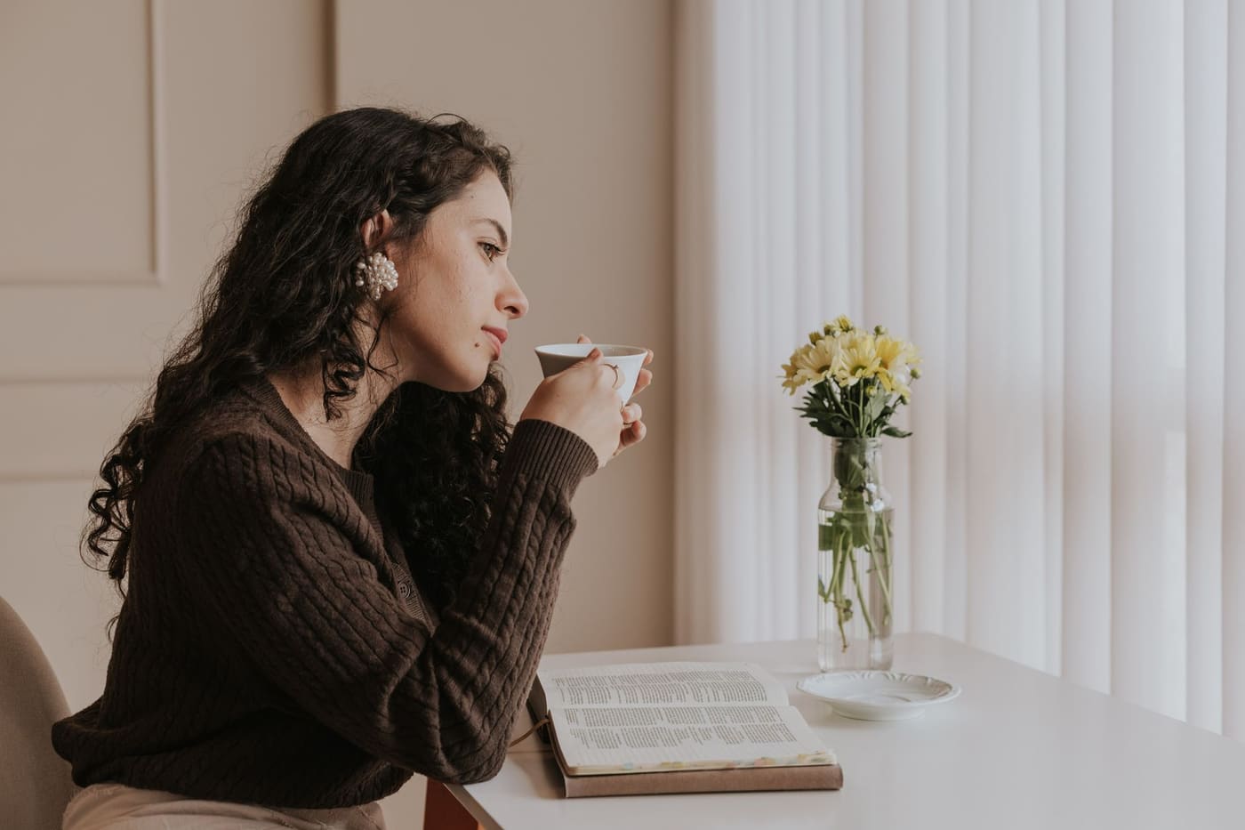 A woman with dark curly hair sipping tea at a table with a book and a small vase of yellow flowers