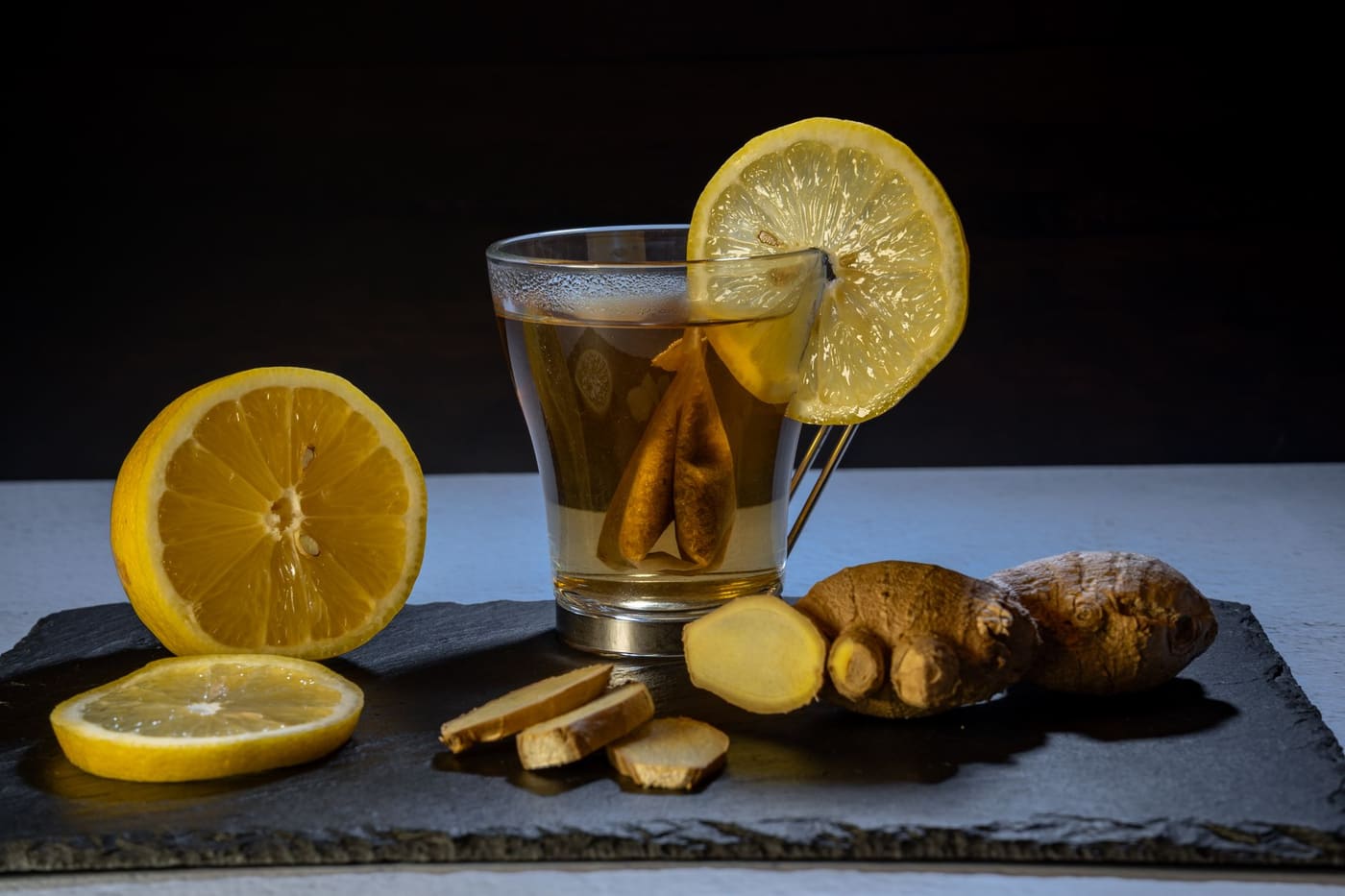 A glass of ginger and lemon tea with lemon slices and ginger root on a slate board