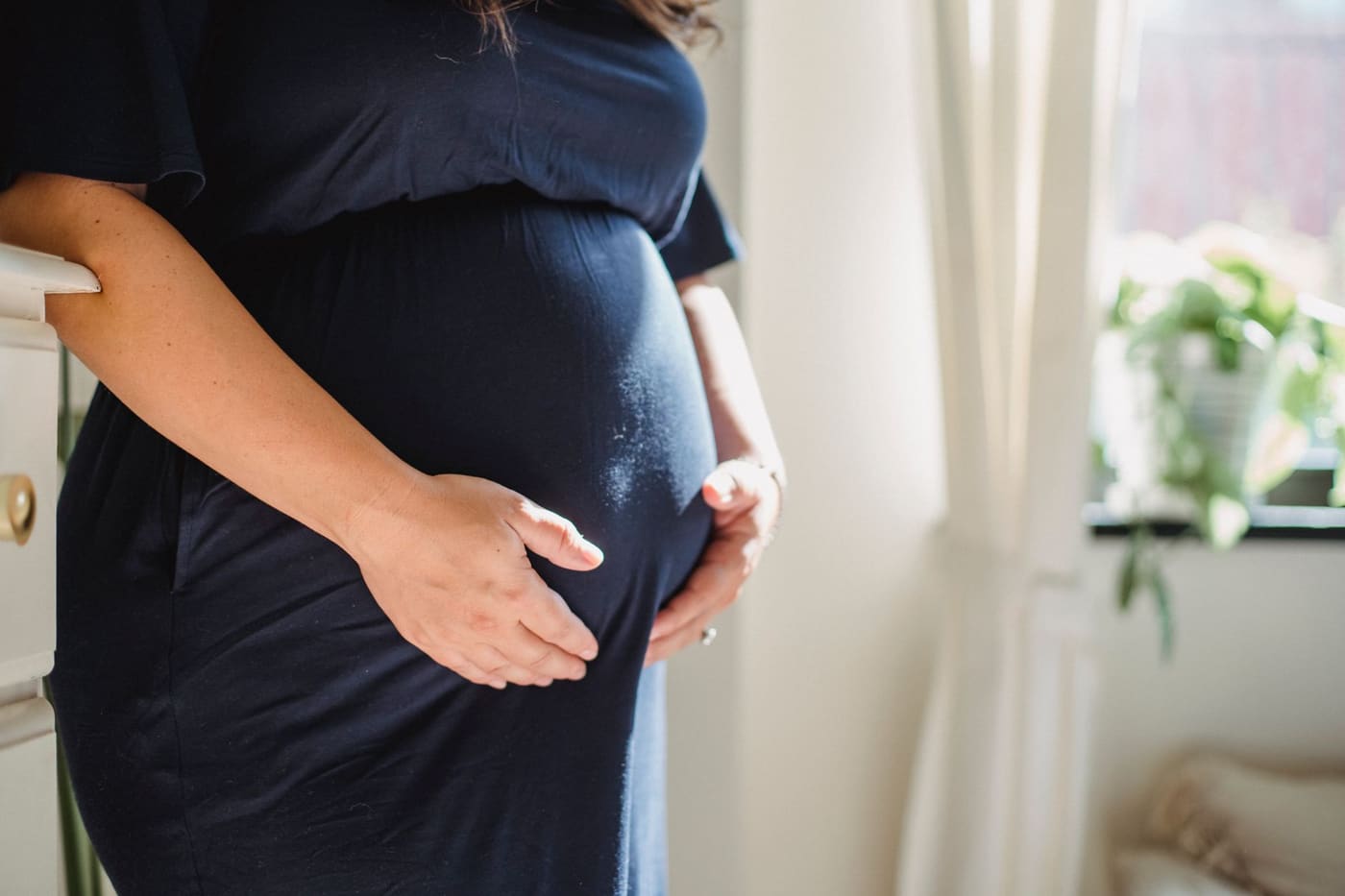 A pregnant woman in a dress with her hands on her belly in a sunlit room