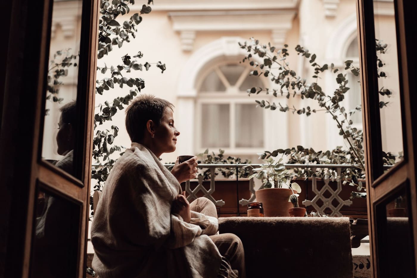 A woman wrapped in a blanket sipping tea on a balcony with plants